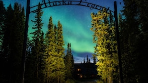 Une enseigne en bois sur laquelle est inscrit « Alaska Legend », placée sur fond de ciel nocturne parsemé d’étoiles et illuminé par les aurores boréales.
