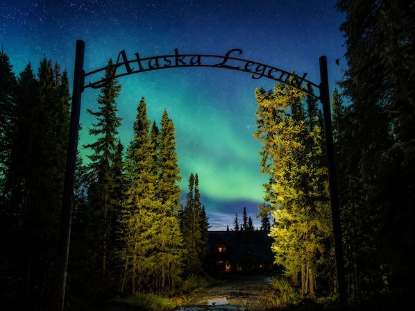 A wooden sign with 'Alaska Legend' written on it, set against a night sky with visible stars and the Northern Lights.