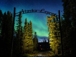 A wooden sign with 'Alaska Legend' written on it, set against a night sky with visible stars and the Northern Lights.