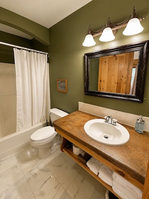 A bathroom with a wooden vanity, a white sink, a mirror, and a shower curtain. - Alaska Legends Kenai River Lodge (Soldotna)