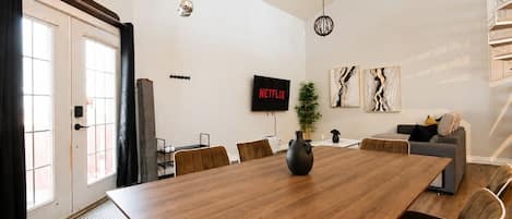 Uma sala de estar moderna com uma grande mesa de jantar de madeira, um sofá e uma TV fixada na parede.