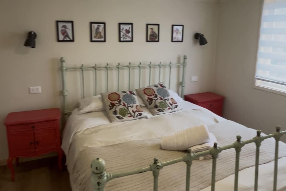 Ein Schlafzimmer mit einem Metallbettgestell, einem roten Nachttisch, einer roten Kommode und einem Fenster mit Jalousien.
