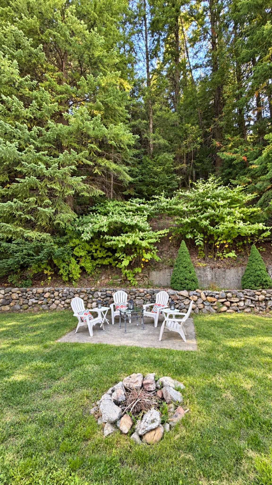 Une terrasse dotée de mobilier d’extérieur et d’un foyer, entourée d’une pelouse, avec un mur de pierres et des arbres en arrière-plan.