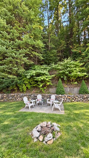 Un patio con mobiliario exterior y un brasero, rodeado de césped, con un muro de piedra y árboles al fondo.