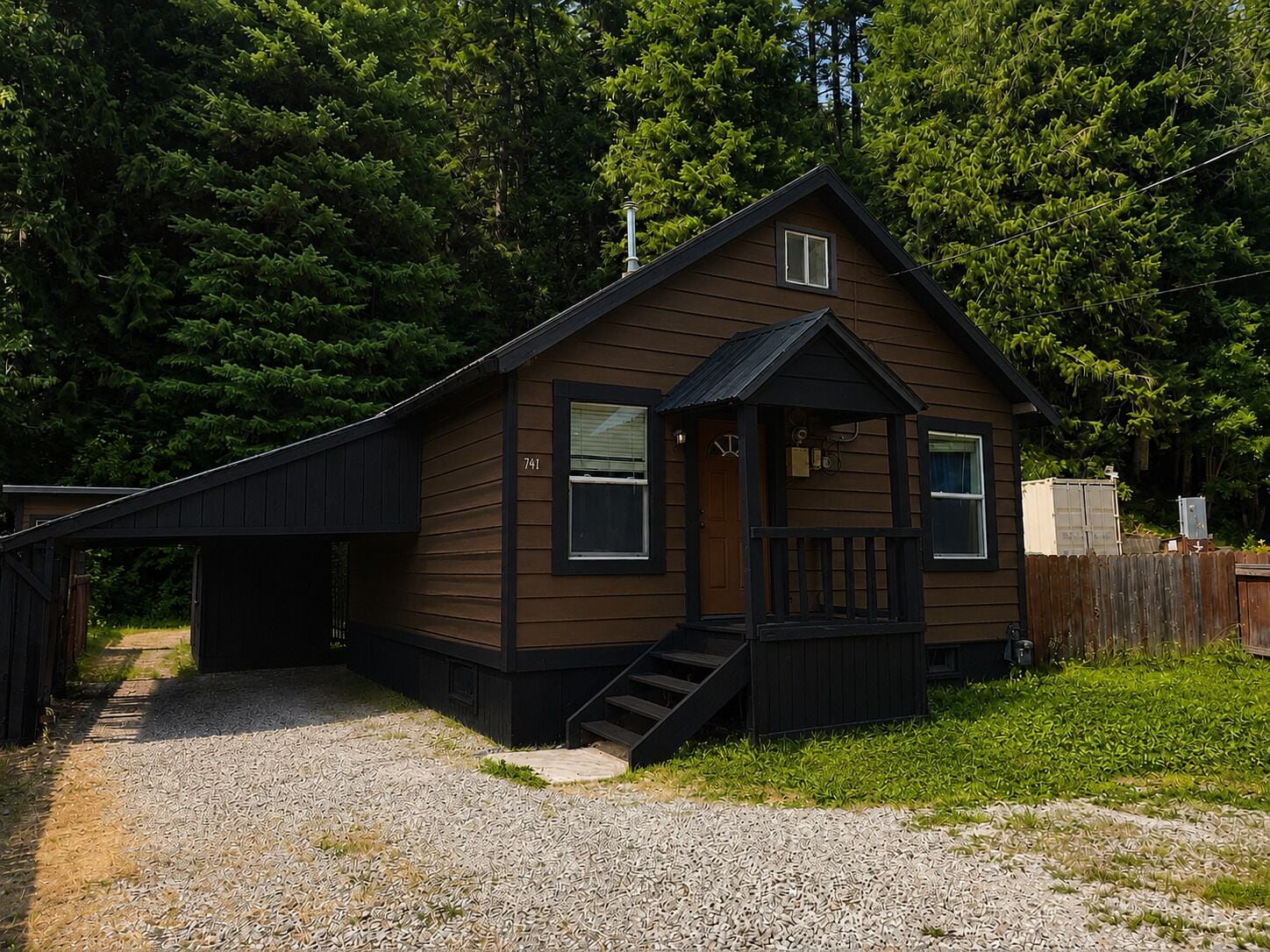 A single-story wooden cabin with a front porch, a small attached garage, and a gravel driveway.