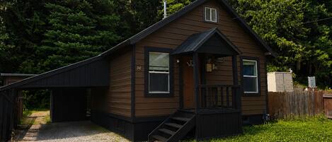 A single-story wooden cabin with a front porch, a small attached garage, and a gravel driveway.