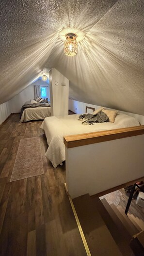 Une chambre mansardée chaleureuse, avec un plafond en pente, un lit doté d’une tête de lit en bois, une petite table et un luminaire décoratif.
