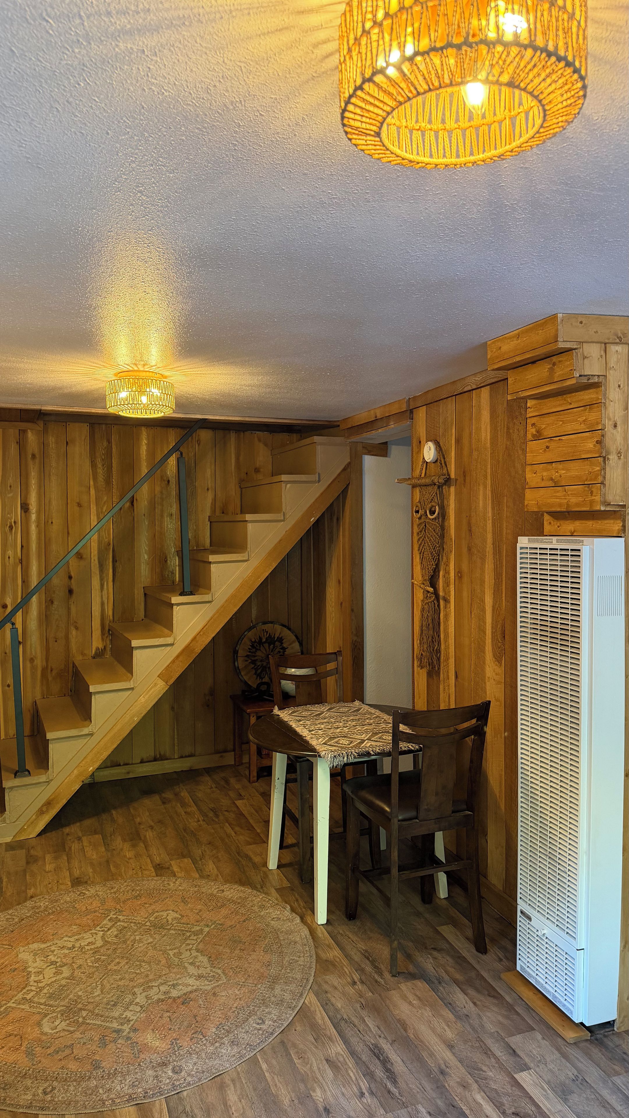 A wooden interior with a staircase, a dining table with chairs, a patterned rug, and a white appliance.