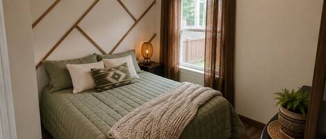 A bedroom with a bed, a bedside lamp, a window with curtains, and a potted plant.