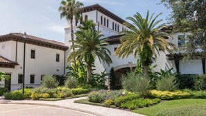 Un edificio de varios pisos con fachada blanca y tejas marrones, rodeado de jardines cuidados y palmeras.
