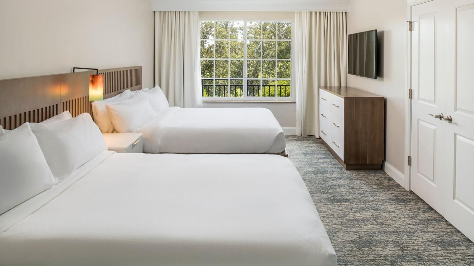 A hotel room with two beds, a TV, a window with curtains, and a dresser.