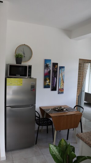 A compact kitchen with a refrigerator, microwave, and a clock on the wall. There is a dining table with chairs and a plant on the floor. - CasaMIA RD: Your Caribbean Getaway. (Río San Juan)
