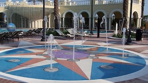 Ein Wasserbrunnen mit mehreren Düsen, umgeben von einem gemusterten Pflasterweg und einem Poolbereich mit Liegestühlen.