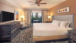 Ein Hotelzimmer mit einem großen Bett, einem Fernseher, einem Schreibtisch, einem Sessel und einem Balkon mit Vorhängen.