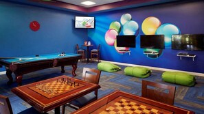 Ein Raum mit Poolbillardtisch, Schachbrett, Billardtisch, grünen Sitzsäcken und einer Wand mit einem farbenfrohen, kugelförmigen Design.