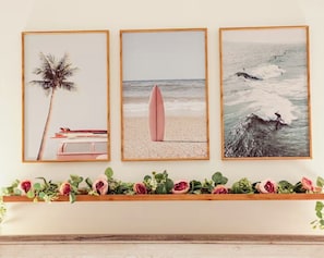 Three framed beach-themed artworks on a wall, a shelf with decorative flowers below, and a wooden shelf above. - King Bed Primary • Spacious Boutique Stay (Homestead)