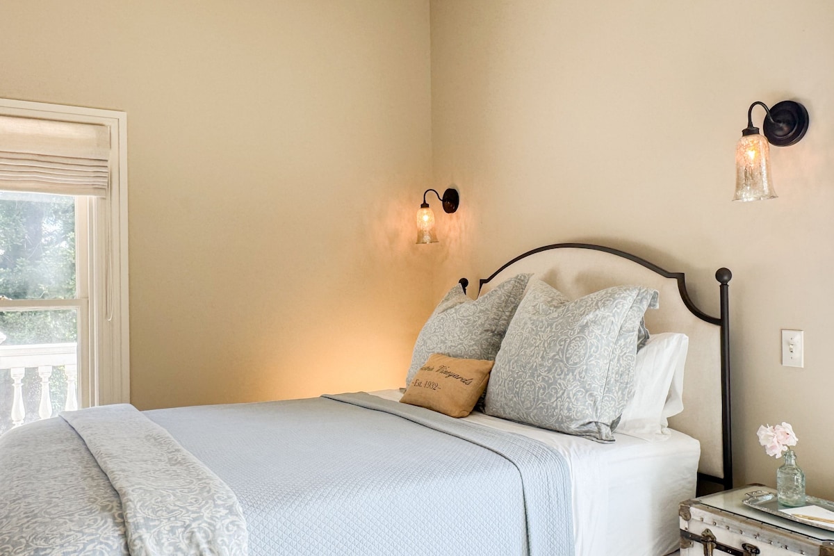 Un dormitorio con cama, mesita de noche, lámpara de pared y ventana con vista a follaje.