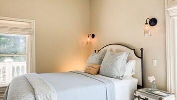 Un dormitorio con cama, mesita de noche, lámpara de pared y ventana con vista a follaje.