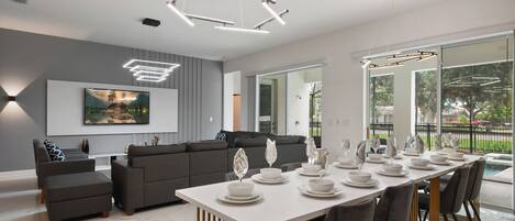 A modern dining area with a long table set for six, a grey sofa, and a large window offering a view of the outdoors.