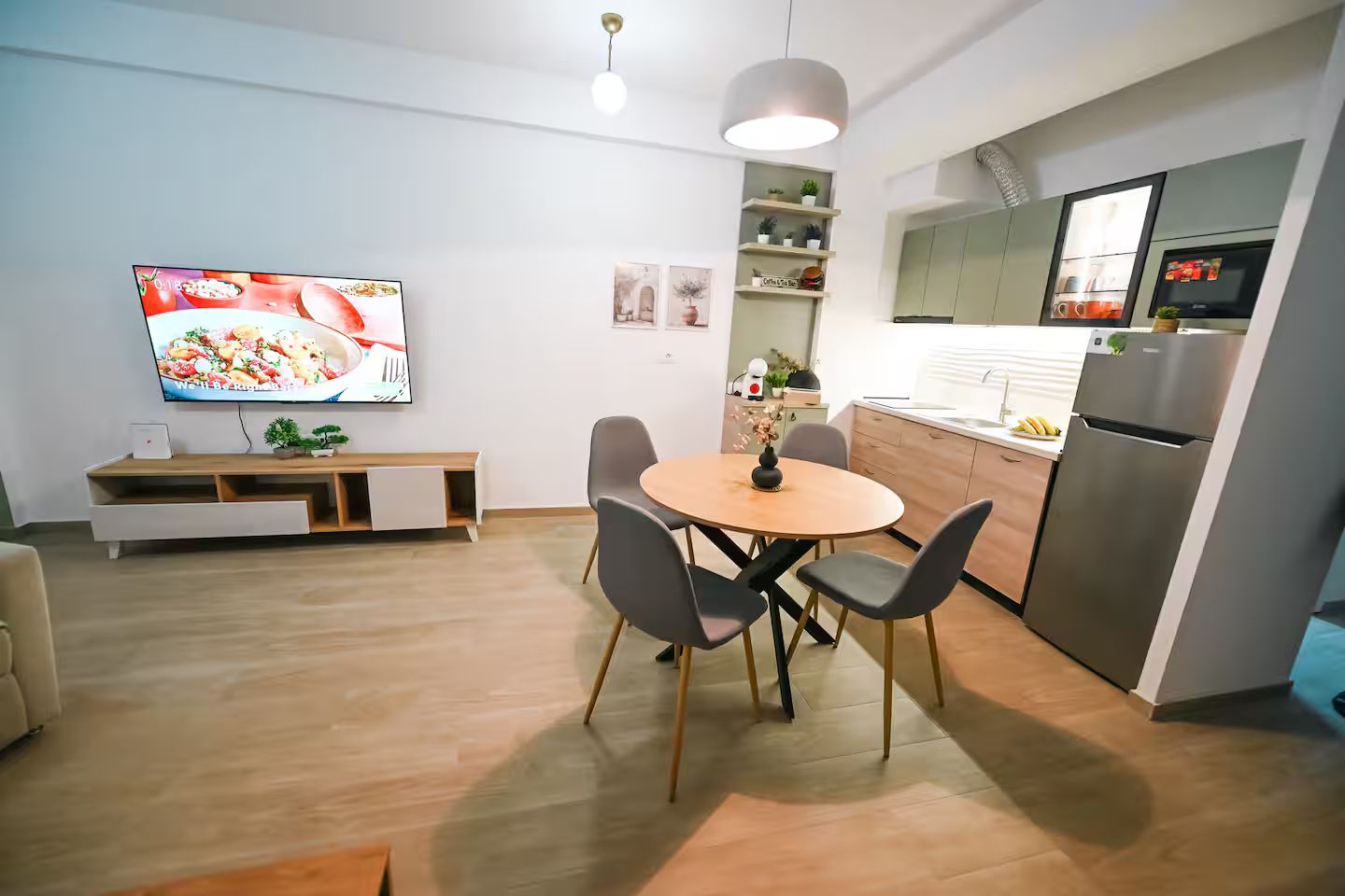 Eine moderne Küche mit Einbauofen und Kühlschrank, ein Essbereich mit rundem Tisch und Stühlen sowie ein an der Wand befestigter Flachbildfernseher.