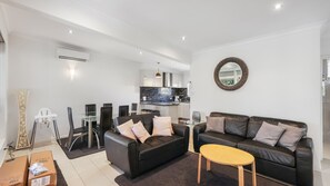 A modern living room with a black leather sofa, a glass dining table, and a kitchen in the background. - PI Holiday Retreat Unit 3 (Cowes)