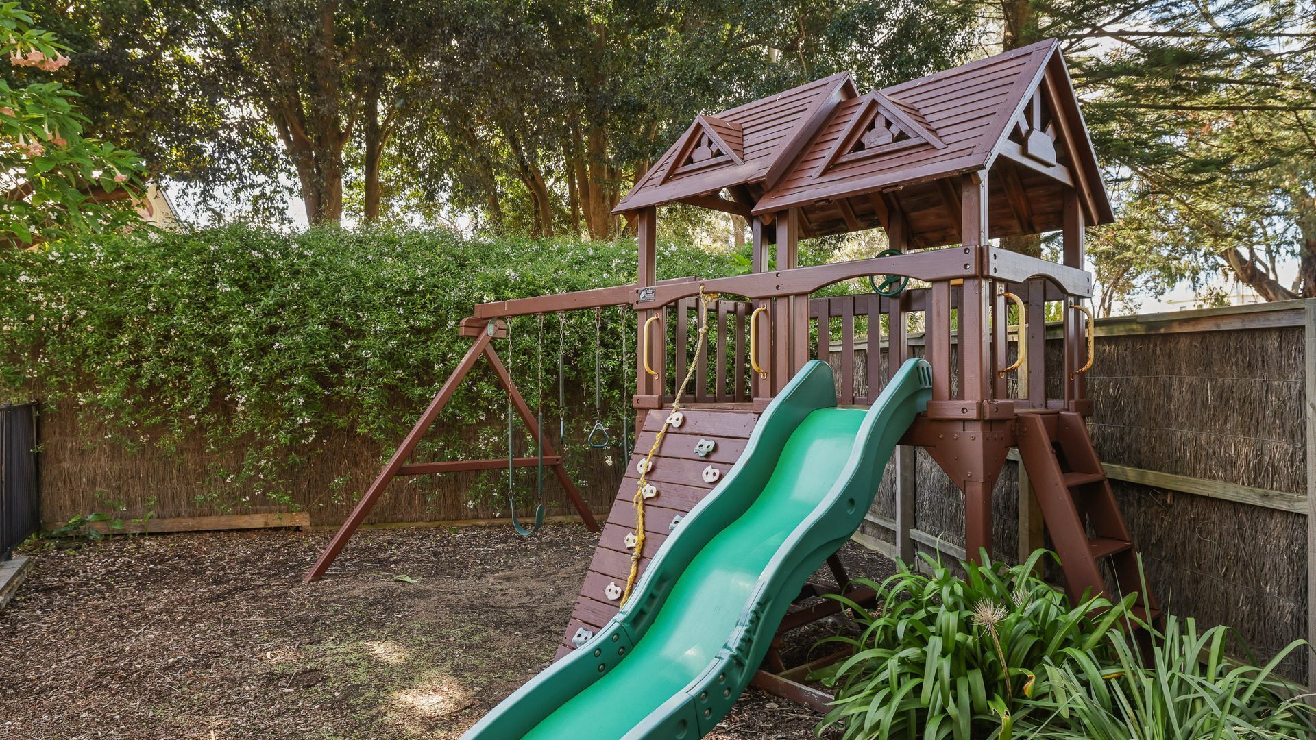 A wooden play structure with a slide and climbing area, surrounded by a fence and greenery.