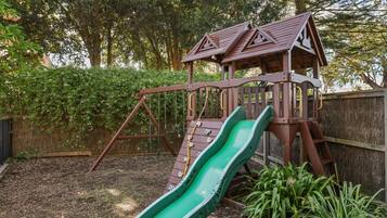 A wooden play structure with a slide and climbing area, surrounded by a fence and greenery.