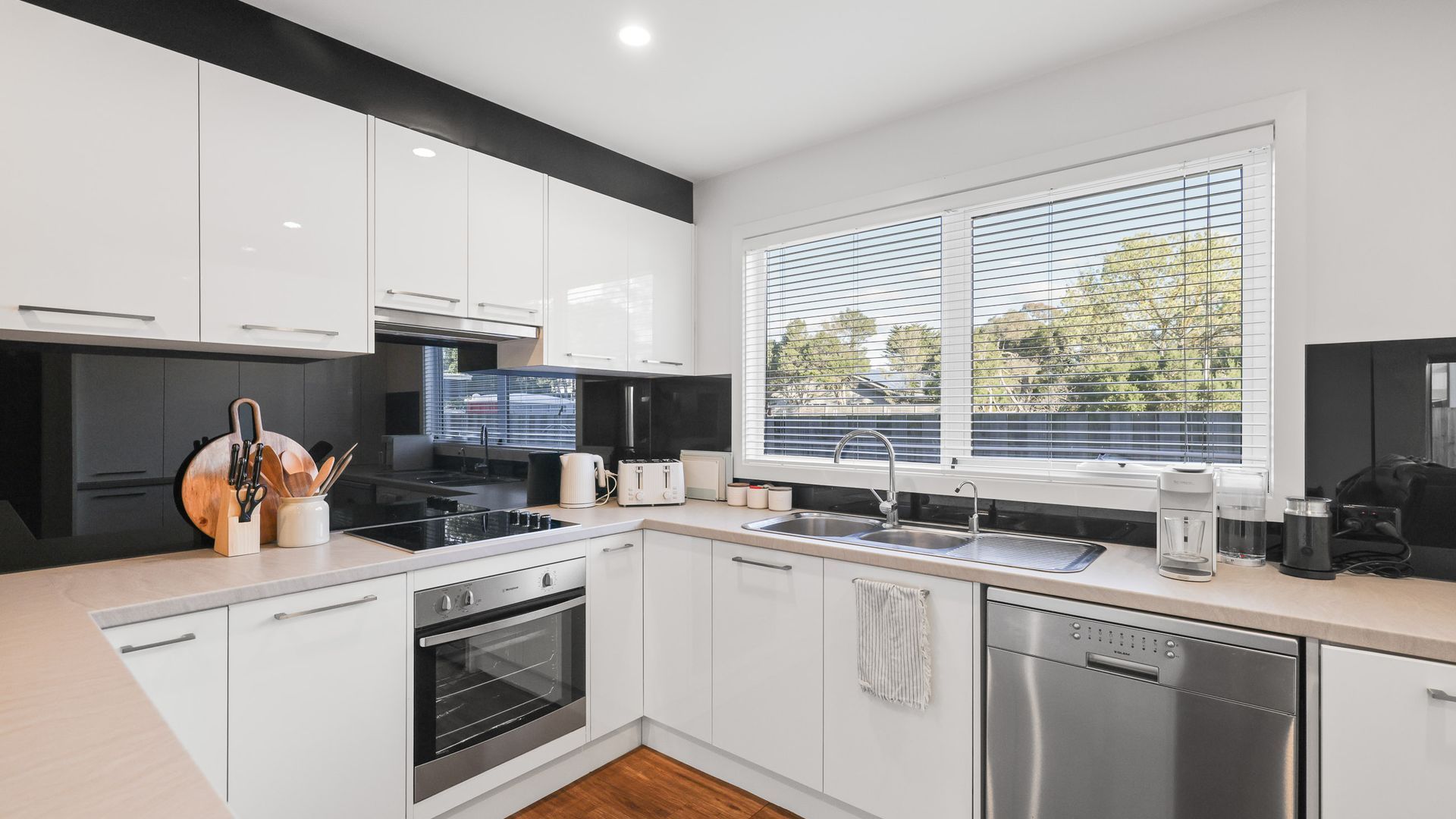 A modern kitchen with white cabinets, a black stove, a dishwasher, and a window with blinds.