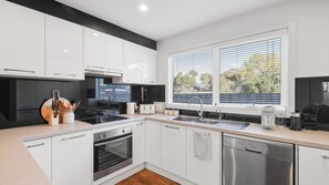 A modern kitchen with white cabinets, a black stove, a dishwasher, and a window with blinds. - Home and Away in Cowes (Cowes)