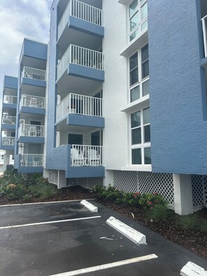Un edifici residencial de diverses plantes amb balcons, una zona d'aparcament i un jardí ple de flors.