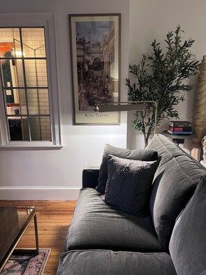 Un salon avec un canapé gris, un tableau encadré au mur, une plante en pot et une fenêtre avec des stores.