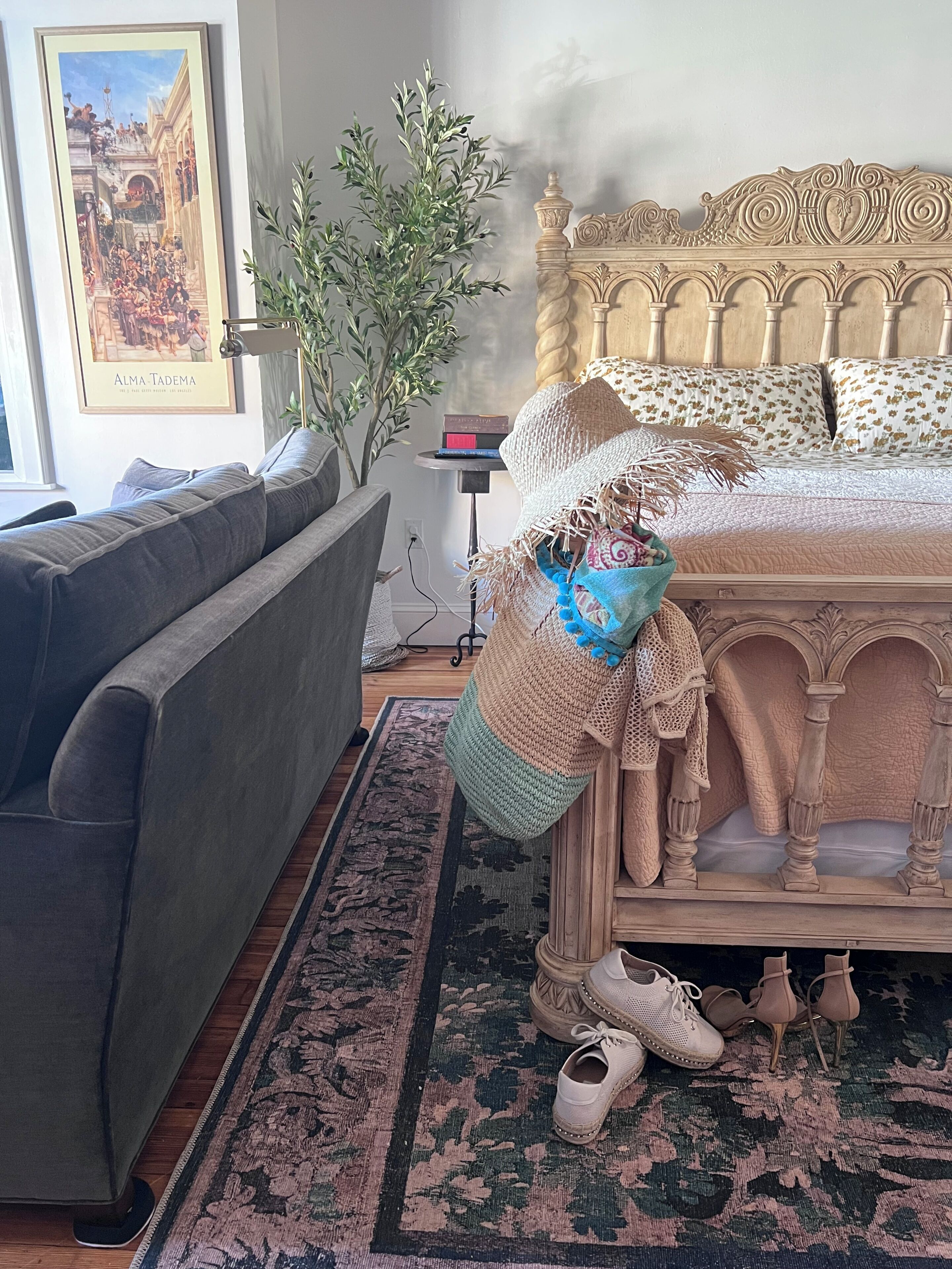 A bedroom with a bed, a grey sofa, a small table with books, a potted plant, and a decorative headboard.
