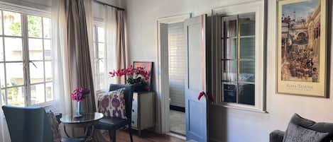 Ein Wohnzimmer mit einer Couch, einem Glastisch, einem Sessel, einem kleinen Tisch mit Büchern, einer Vase mit Blumen und einer Tür zu einem anderen Raum.