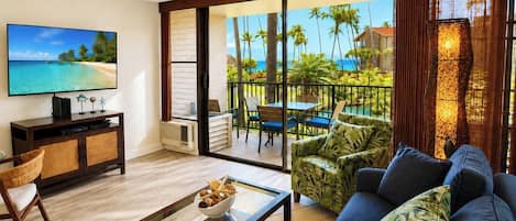 A living room with a television, a coffee table, and a sofa. There is a balcony with a view of palm trees and the ocean.