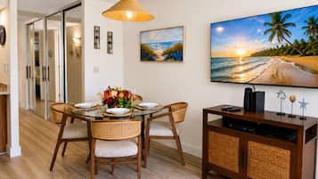 Un comedor con una mesa de vidrio, sillas de mimbre, una lámpara de techo tejida, un cuadro de una playa y un televisor de pantalla plana sobre un mueble de madera.