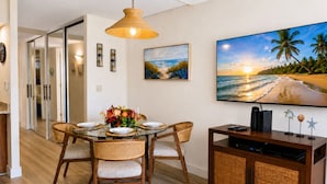 Un'area pranzo con tavolo in vetro, sedie in vimini, lampada a soffitto intrecciata, un quadro raffigurante una spiaggia e un televisore a schermo piatto su un mobile in legno.