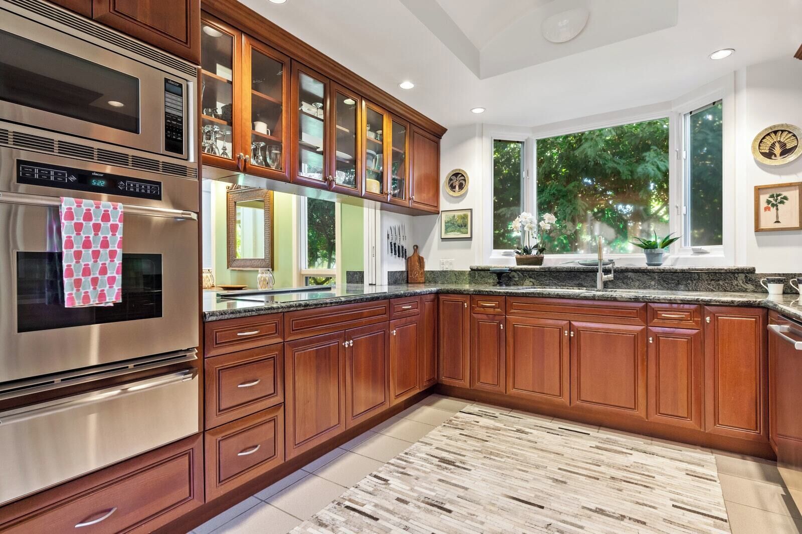 Cocina con armarios de madera, electrodomésticos de acero inoxidable, encimera de granito y un amplio ventanal con vistas a zonas verdes.