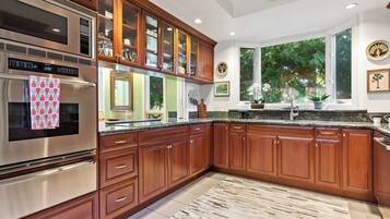 Cocina con armarios de madera, electrodomésticos de acero inoxidable, encimera de granito y un amplio ventanal con vistas a zonas verdes.