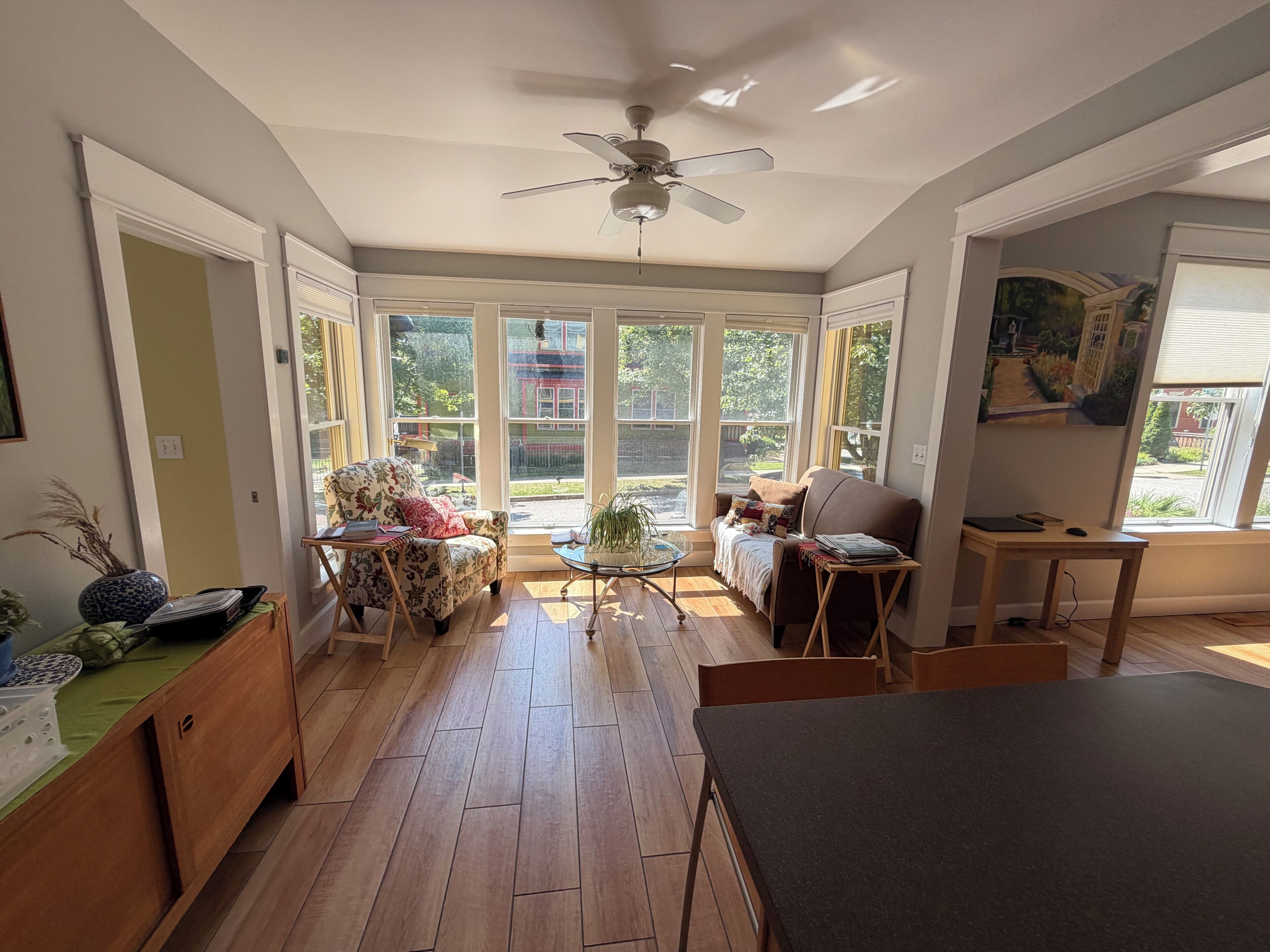 Une pièce baignée de lumière, avec un ventilateur de plafond, un canapé, une table à manger et une vue sur un jardin, visible à travers de grandes fenêtres.