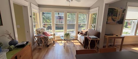 A sunlit room with a ceiling fan, a sofa, a dining table, and a view of a garden through large windows.