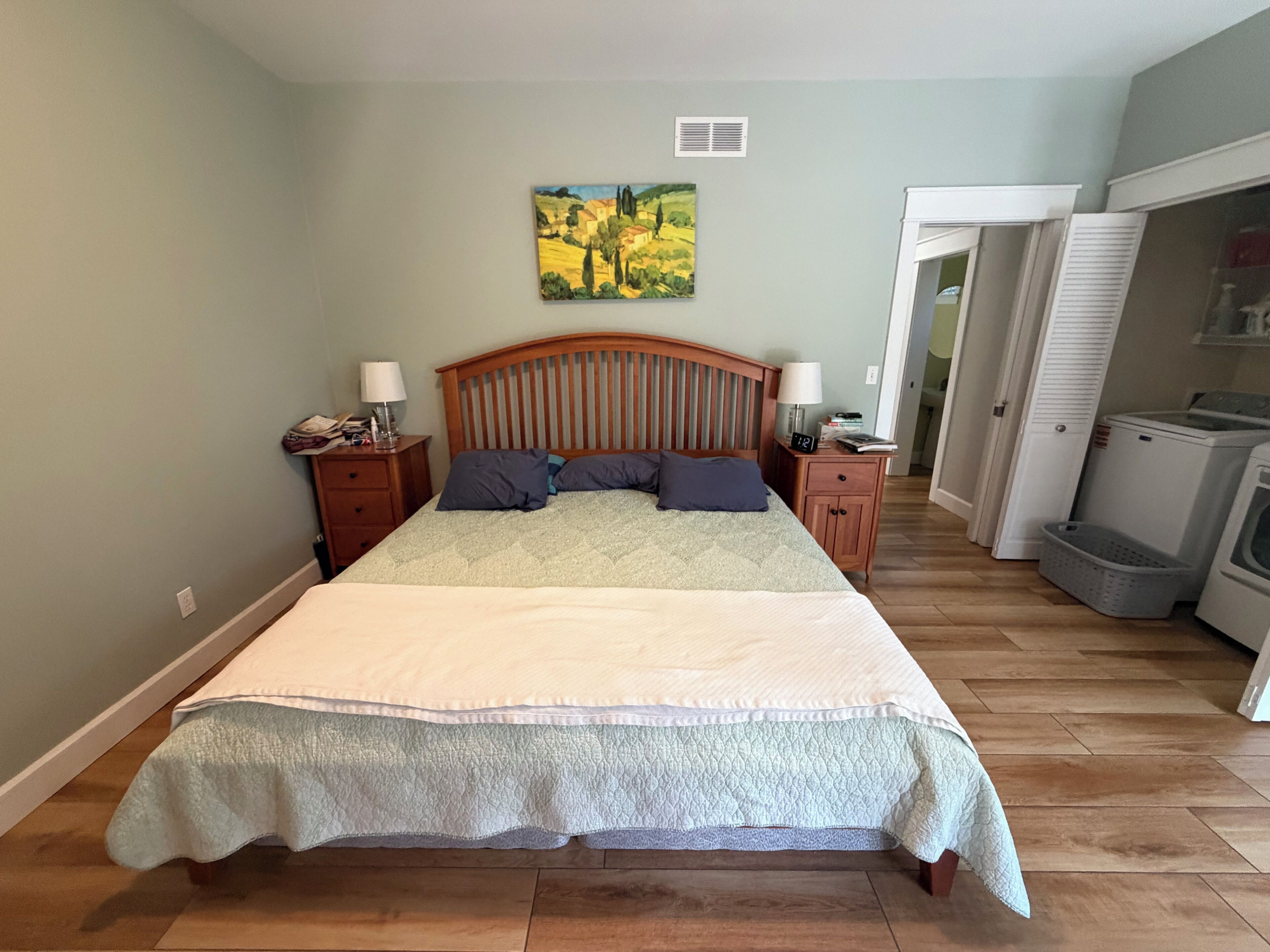 A bedroom with a large bed, wooden headboard, two bedside tables with lamps, a painting on the wall, and a laundry area with appliances.