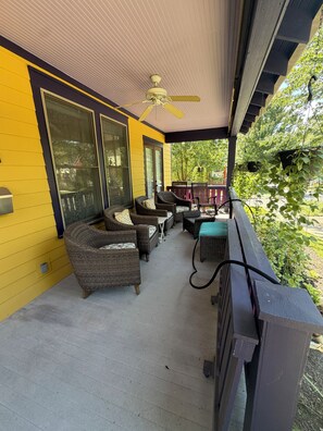 A covered porch with wicker furniture, a ceiling fan, and a view of greenery.