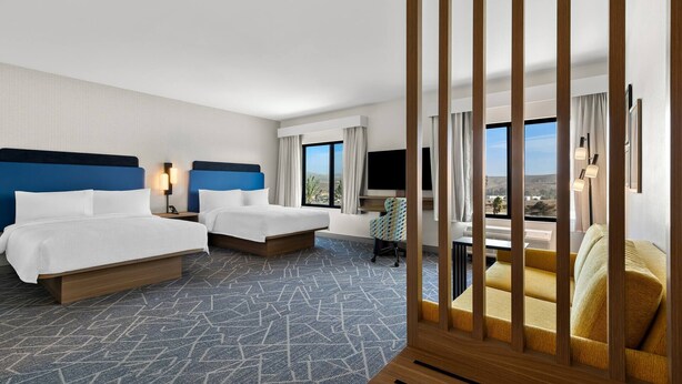 A hotel room with two beds, a desk, a chair, and a large window offering a view.
