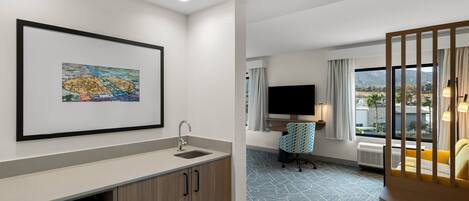 A modern hotel room with a kitchenette, a flat-screen TV, a comfortable seating area, and a large window offering a view.