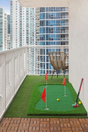 Ein kleiner Indoor-Putting-Grün mit einem Stuhl und Golfschlägern, umgeben von einer weißen Brüstung und Blick auf die Stadt.