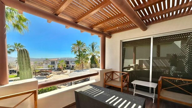 Un balcón con una pérgola de madera, una mesa y sillas, con vista a un paisaje urbano con palmeras y edificios.