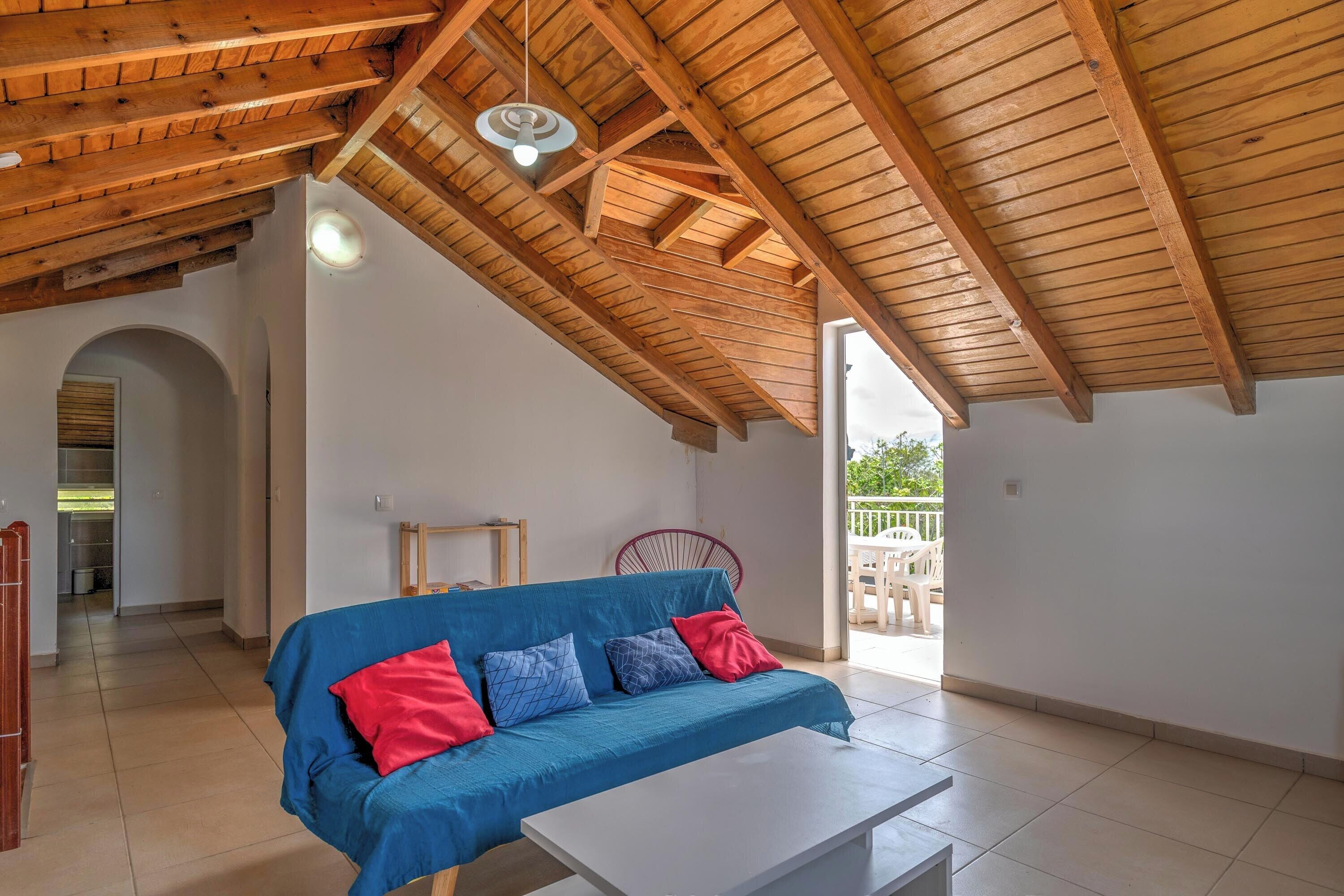 A spacious room with a high wooden ceiling, a blue sofa with red and blue cushions, a white table, and a view of the outside.