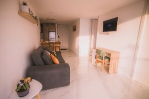 Un saló modern amb un sofà gris, una taula petita amb una planta, un escriptori de fusta i un televisor fixat a la paret.