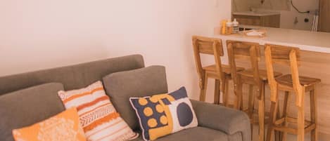 A living room with a grey sofa, wooden chairs, a small kitchen area, and a clock on the wall.