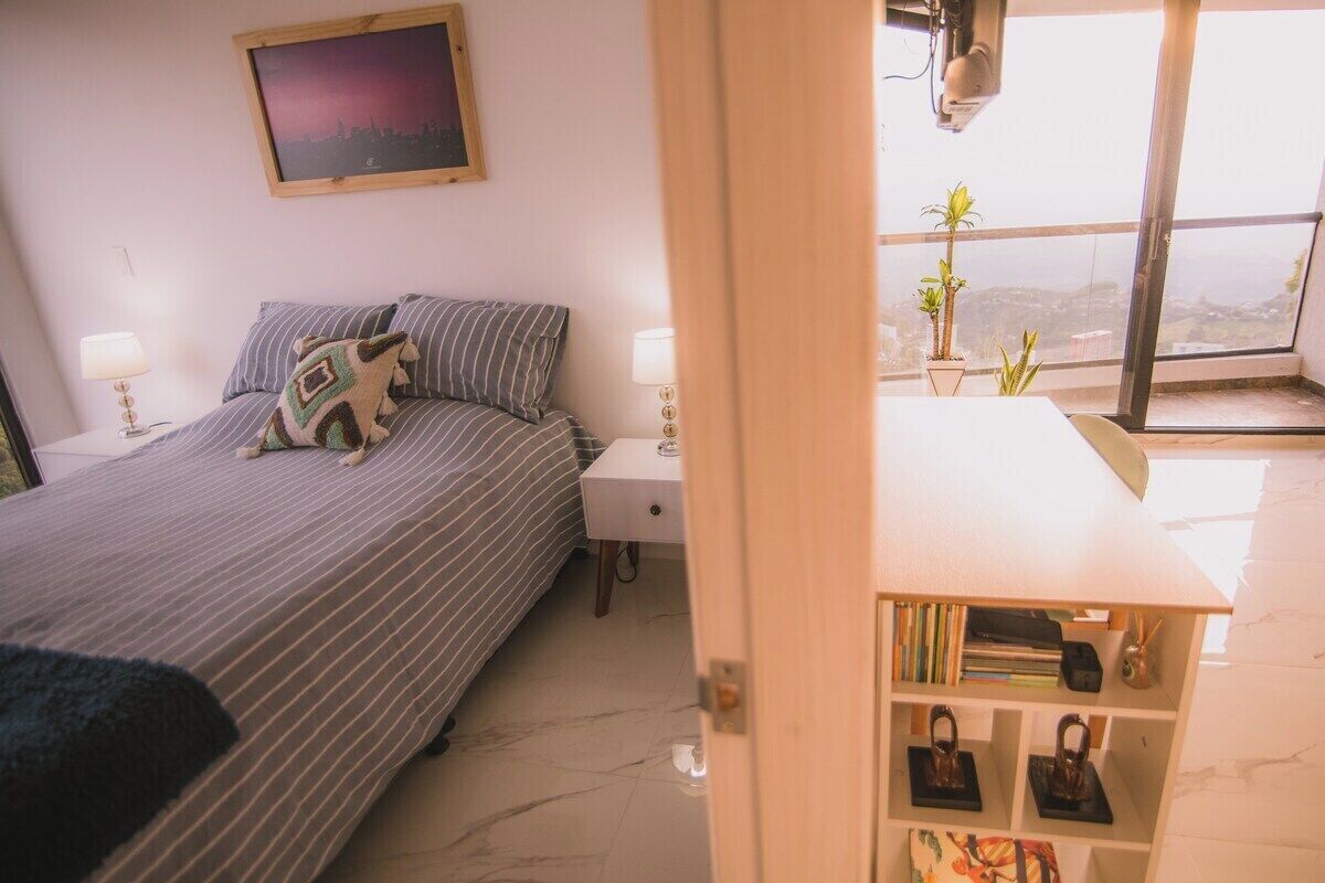 Un dormitorio con cama, mesitas de noche, estantería y ventana con vistas.